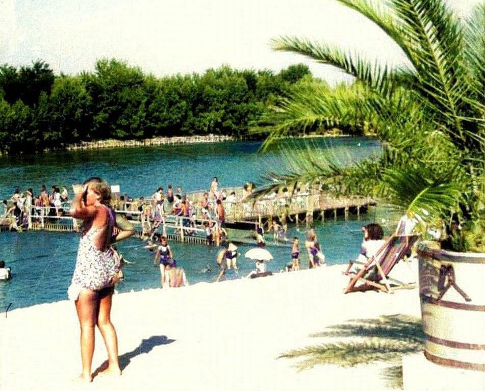 Kupači na pješčanoj plaži i drvenom pontonu Garnizonskog kupališta na Dravi, u prvom planu posuda s palmom.